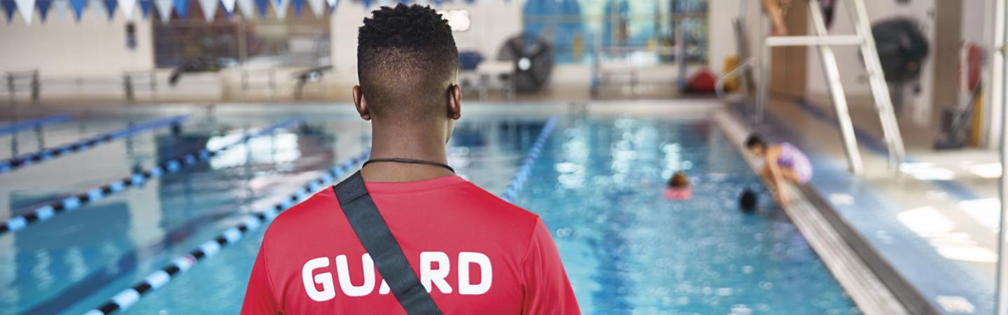 A lifeguard faces a pool and his red "lifeguard" shirt is in the forefront